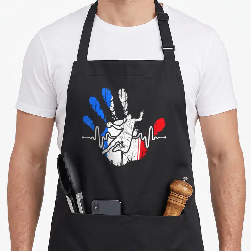 Tablier de cuisine noir avec illustration d'un handballeur sautant devant une empreinte de main aux couleurs du drapeau français et une ligne d'électrocardiogramme.