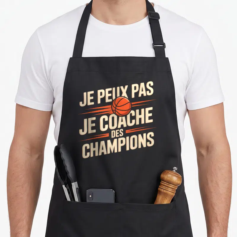 Tablier de cuisine noir avec l'inscription Je peux pas je coache des champions et un ballon de basket orange au centre du design typographique.