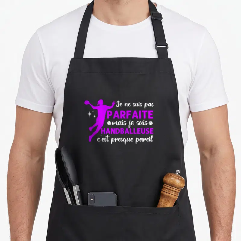 Tablier de cuisine noir avec une silhouette violette d'une handballeuse et un texte blanc et rose affirmant la perfection du sport.