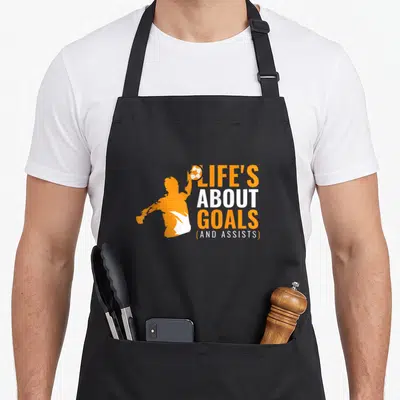 Tablier de cuisine noir avec silhouette orange d'un joueur de sport et texte Life's About Goals And Assists en blanc et orange.