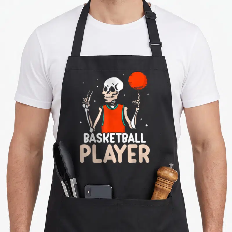 Tablier de cuisine noir avec squelette en maillot orange faisant tourner un ballon rouge, texte Basketball Player en bas.