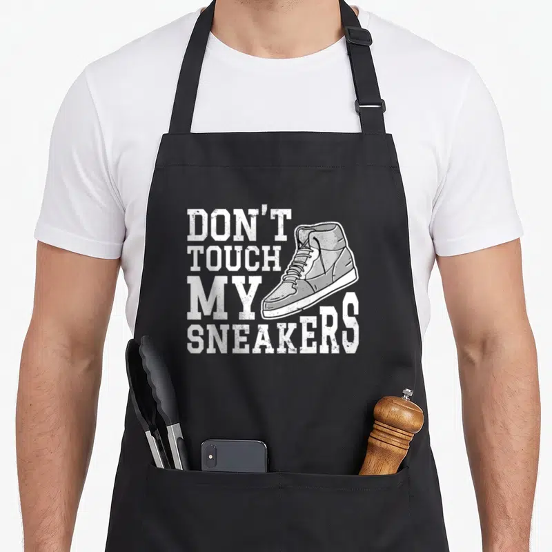 Tablier de cuisine noir avec illustration de sneaker blanche et texte Don't Touch My Sneakers en typographie sport.