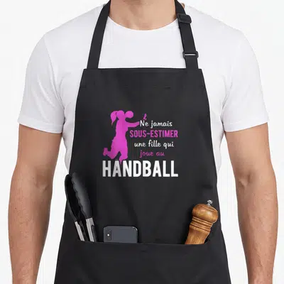 Tablier de cuisine noir avec silhouette fuchsia d'une joueuse de handball et texte blanc et roseant sur le devant.