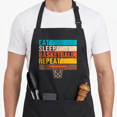 Tablier de cuisine noir avec le texte Eat Sleep Basketball Repeat et un dessin de cerceau de panier en dessous.
