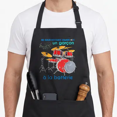 Tablier de cuisine noir avec illustration d'un kit de batterie rouge, cymbales dorées et texte bleu sur fond uni.