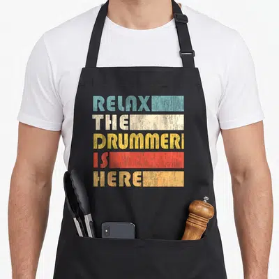 Tablier de cuisine noir avec le texte Relax The Drummer Is Here en lettres colorées style rétro sur fond uni.