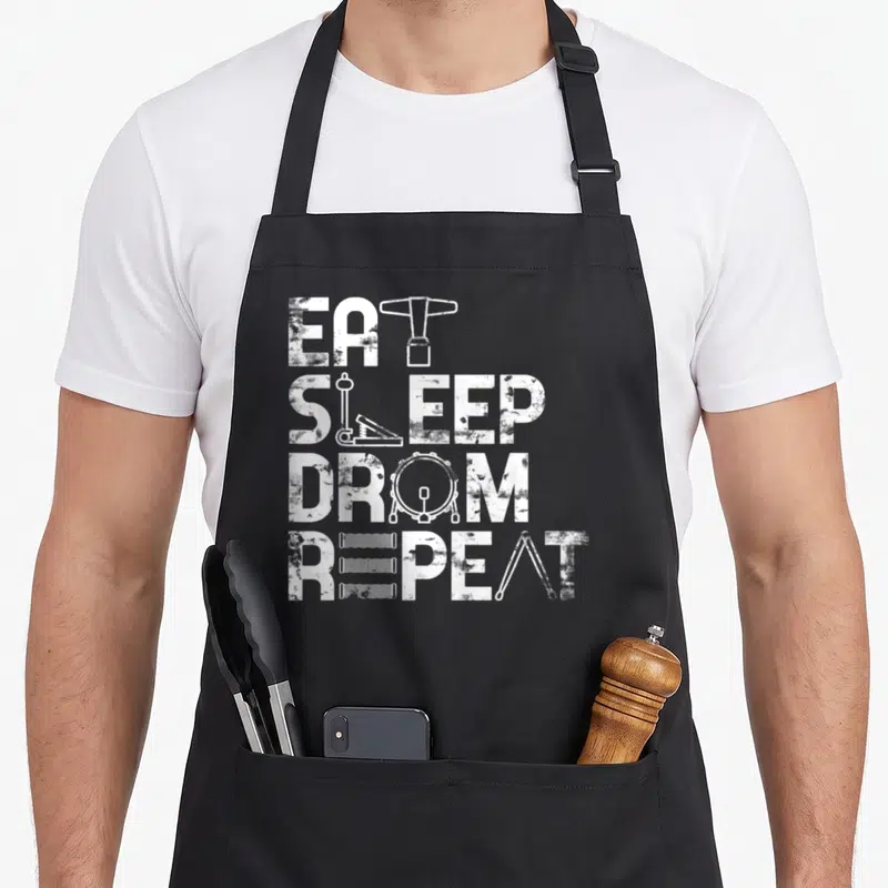 Tablier de cuisine noir avec le motifEat Sleep Drum Repeat intégrant des instruments de percussion en blanc.