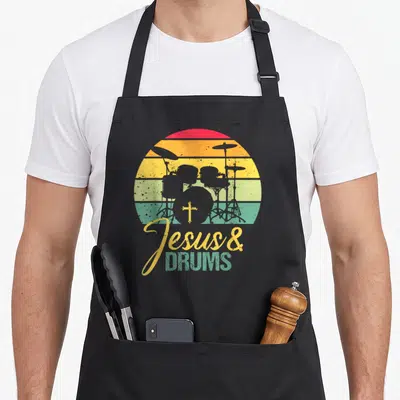 Tablier de cuisine noir avec illustration vintage de kit de drums, croix et texte Jesus & Drums sur fond de soleil arc-en-ciel.