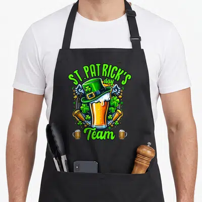 Tablier de cuisine noir avec illustration colorée d'un verre de bière surmonté d'un chapeau vert et du texte St. Patrick's Day Team.