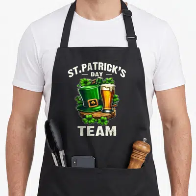 Tablier de cuisine noir avec illustration d'un chapeau vert, d'une bière et de trèfles sur un support en bois.