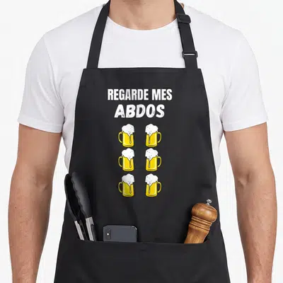 Tablier de cuisine noir avec texte blanc Regarde mes abdos et six mugs de bière jaune alignés en deux colonnes.