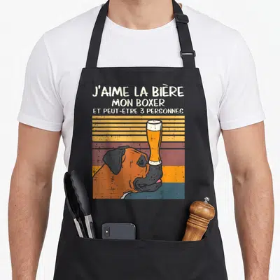 Tablier de cuisine noir avec illustration d'un chien boxer tenant une coupe de bière sur le nez et texte humoristique en lettres blanches sur fond rayé vintage.