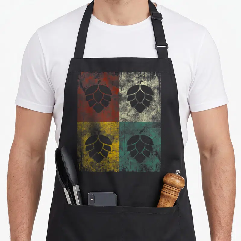 Tablier de cuisine noir avec quatre feuilles de houblon stylisées sur fonds texturés rouge, beige, jaune et vert.