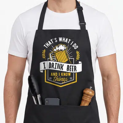 Tablier de cuisine noir avec illustration d'une grande tasse de bière et texte humoristique en blanc et jaune.