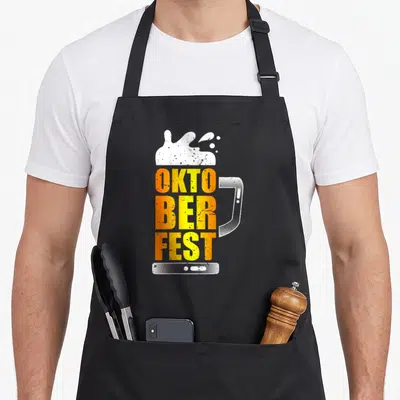 Tablier de cuisine noir avec un grand mug stylisé orange et blanc affichant le texte OKTOBERFEST sous une mousse blanche.