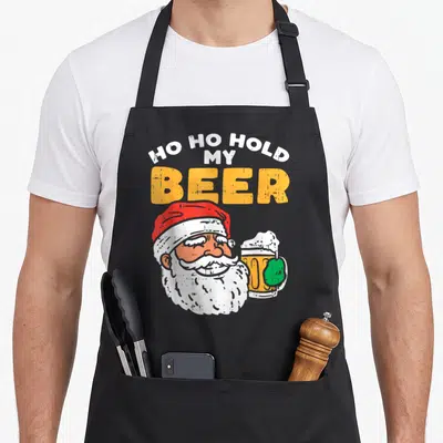 Tablier de cuisine noir avec illustration d'un Père Noël tenant une bière et le texte Ho ho hold my beer en orange et blanc.