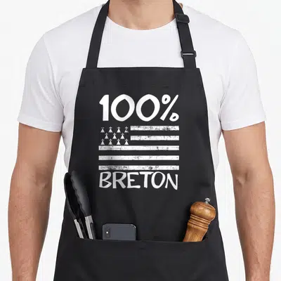 Tablier de cuisine noir avec inscription 100 % BRETON et motifs de coiffes traditionnelles blanches sur fond texturé.