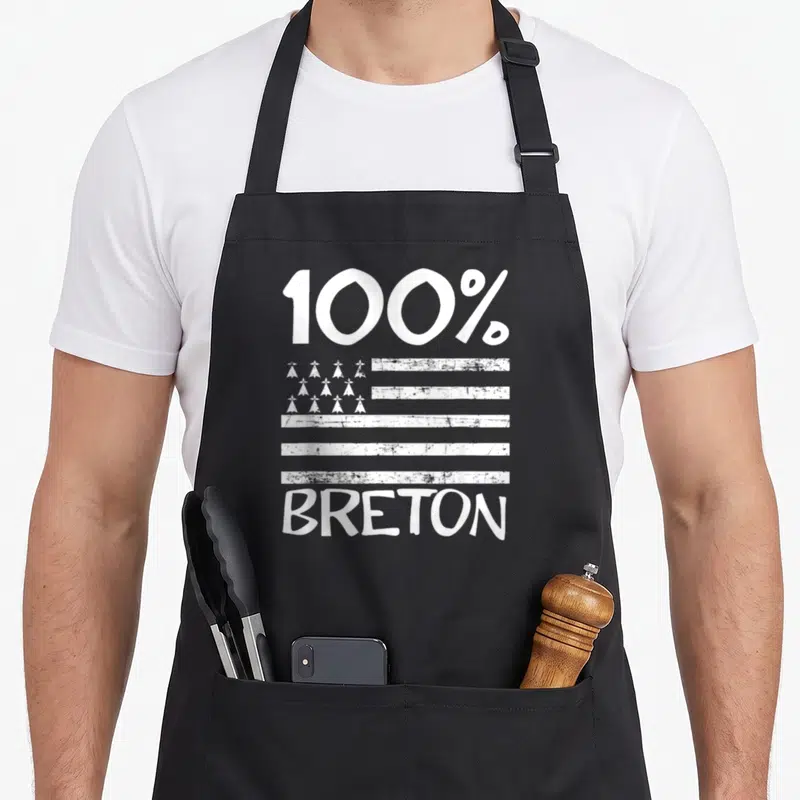 Tablier de cuisine noir avec inscription 100 % BRETON et motifs de coiffes traditionnelles blanches sur fond texturé.