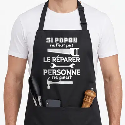Tablier de cuisine noir avec inscription blanche et outils comme une scie, une clé et un marteau.