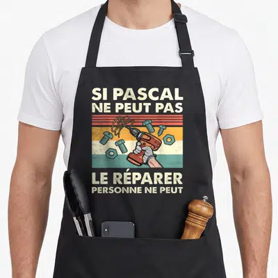 Tablier de cuisine noir avec une illustration de perceuse orange, de vis bleues et le texte Si Pascal ne peut pas le réparer personne ne peut.