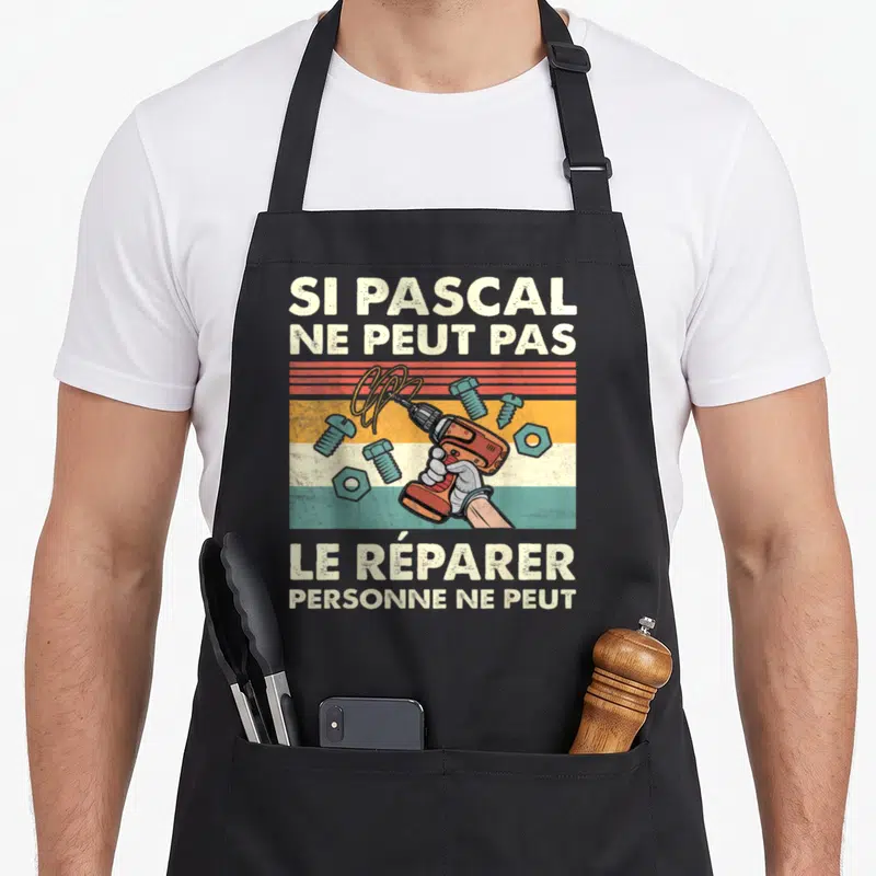 Tablier de cuisine noir avec une illustration de perceuse orange, de vis bleues et le texte Si Pascal ne peut pas le réparer personne ne peut.