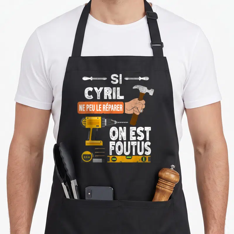 Tablier de cuisine noir avec une perceuse jaune, un marteau et l'inscription Si Cyril ne peut le réparer on est foutus.