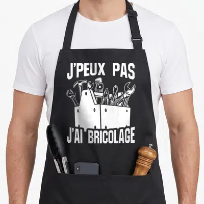 Tablier de cuisine noir avec une illustration blanche d'une boîte à outils et le texte J'peux pas j'ai bricolage en haut et en bas.
