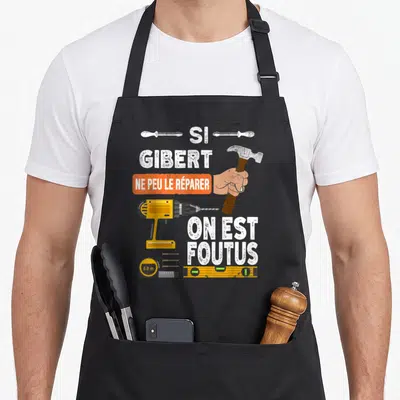Tablier de cuisine noir avec illustration de marteau, perceuse jaune et texte humoristique sur fond gris uni.