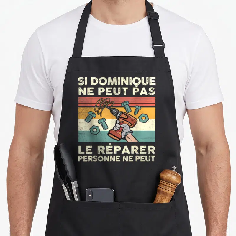Tablier de cuisine noir avec illustration vintage d'une main tenant une perceuse orange sur fond de bandes colorées et vis.