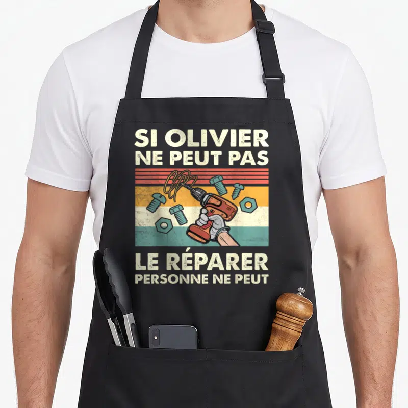 Tablier de cuisine noir avec une illustration vintage d'une main tenant une perceuse entourée de vis et de boulons sur fond multicolore.