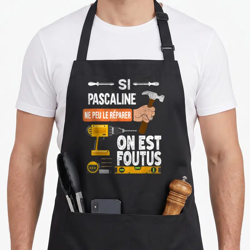 Tablier de cuisine noir avec illustration d'une perceuse, d'un marteau et le texte humoristique sur fond gris uni.