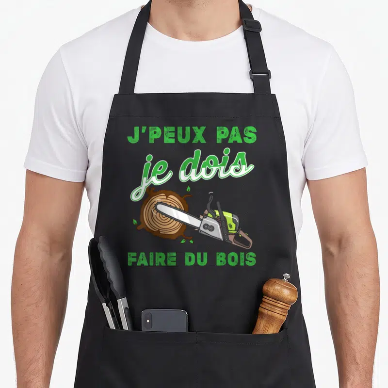 Tablier de cuisine noir avec inscription verte J'peux pas je dois faire du bois et une tronçonneuse sur un tronc d'arbre.