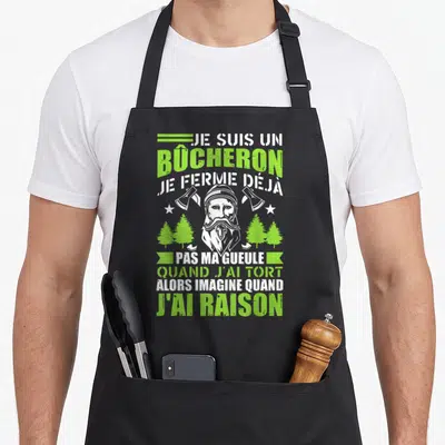 Tablier de cuisine noir avec illustration d'un bûcheron barbu, deux haches et des pins verts. Texte blanc et vert sur fond noir.