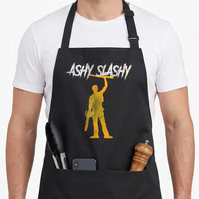 Tablier de cuisine noir avec silhouette orange tenant une tronçonneuse et texte Aschy Slash My en haut.