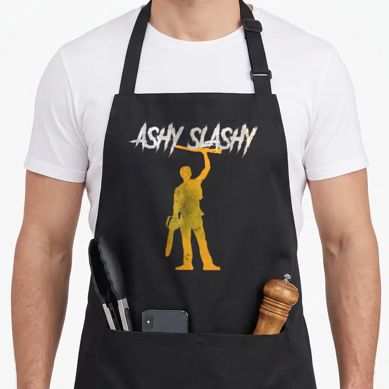 Tablier de cuisine noir avec silhouette orange tenant une tronçonneuse et texte Aschy Slash My en haut.