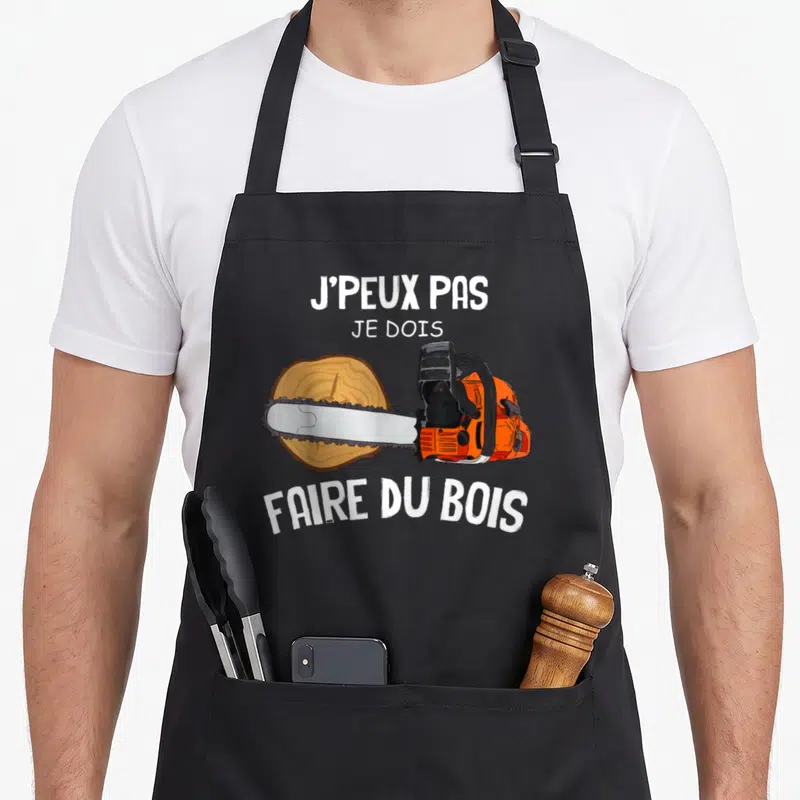 Tablier de cuisine noir avec une tronçonneuse orange traversant un rondin et le texte J'peux pas je dois faire du bois en blanc.