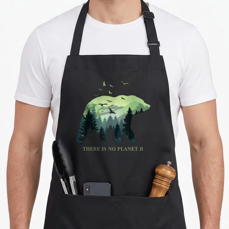 Tablier de cuisine noir avec silhouette d'ours contenant une forêt et des oiseaux en vol au-dessus.