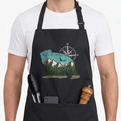 Tablier de cuisine noir avec silhouette d'ours contenant montagnes, forêts et boussole directionnelle.