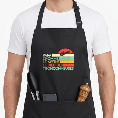 Tablier de cuisine noir avec inscription Papa l'homme le mythe le dieu des tronçonneuses et silhouette de tronçonneuse sur fond de soleil rayé.