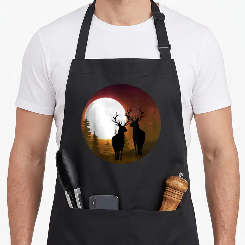 Tablier de cuisine noir avec illustration circulaire de deux cerfs silhouettés devant une lune pleine dans un bois d'hiver.