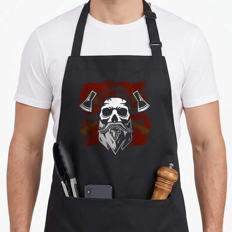 Tablier de cuisine noir avec tête de mort barbue et deux haches croisées sur fond rouge éclaboussé.