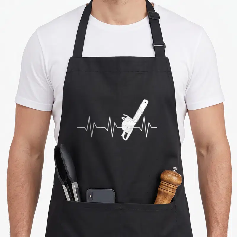 Tablier de cuisine noir avec tracé d'électrocardiogramme blanc intégrant une tronçonneuse en guise de battement de cœur