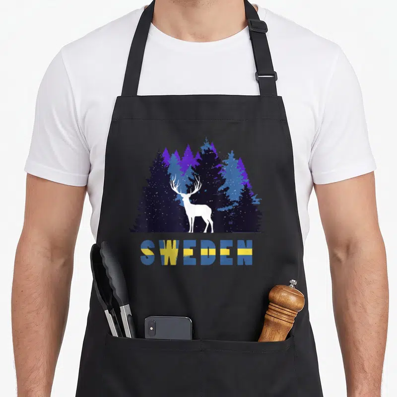 Tablier de cuisine noir avec silhouette d'orignal blanc devant une forêt bleue et le mot SWEDEN aux couleurs du drapeau.
