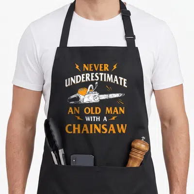 Tablier de cuisine noir avec une tronçonneuse orange et le texte Ne sous-estimez jamais un vieil homme avec une tronçonneuse.