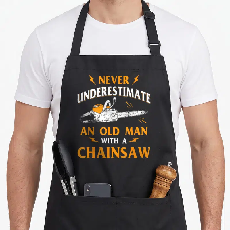 Tablier de cuisine noir avec une tronçonneuse orange et le texte Ne sous-estimez jamais un vieil homme avec une tronçonneuse.