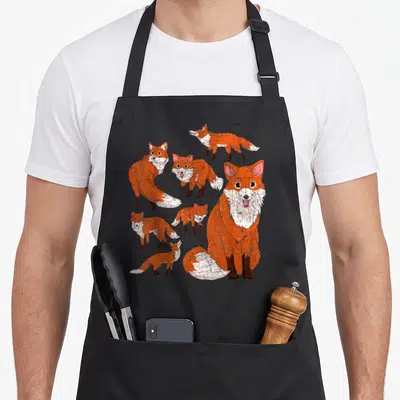 Tablier de cuisine noir avec plusieurs illustrations de renards roux souriants et expressifs dans un style dessin animé.