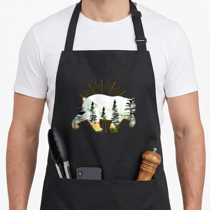 Tablier de cuisine noir avec silhouette d'ours contenant une illustration de forêts et rayons dorés.