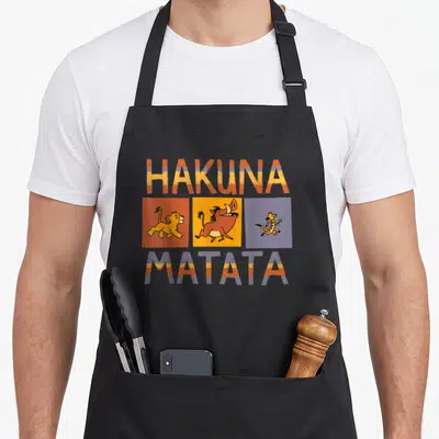 Tablier de cuisine noir avec les mots Hakuna Matata et les personnages Simba, Pumbaa et Timon dans des carrés orange, jaune et violet.