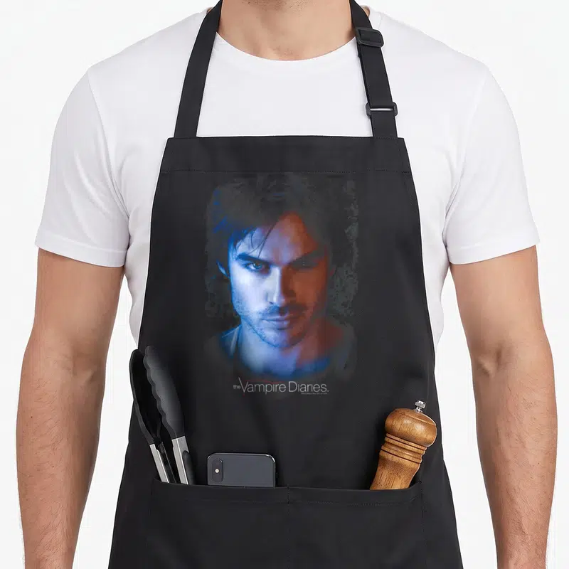 Tablier de cuisine noir avec portrait artistique de Damon Salvatore aux couleurs bleu et rouge, sous le logo The Vampire Diaries.