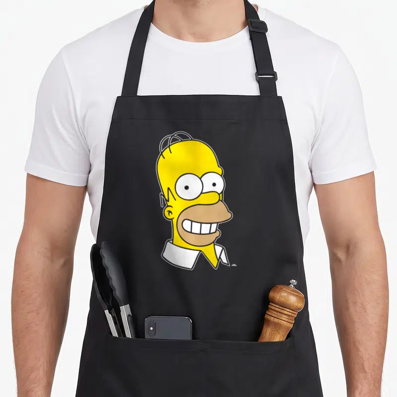 Tablier de cuisine noir avec un grand visage jaune de Homer Simpson affichant un large sourire et trois cheveux sur le crâne.